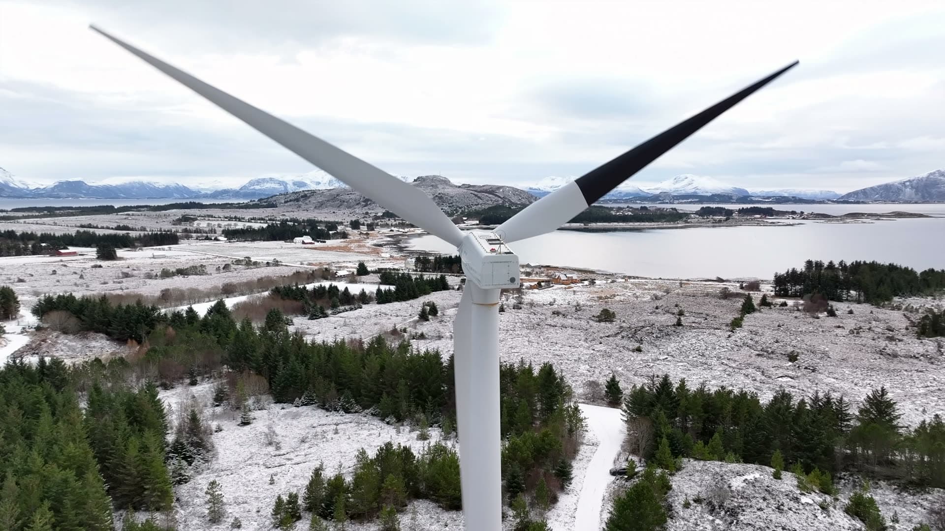 Rotating wind turbine above snowy ground and coastal woodland, Harøy