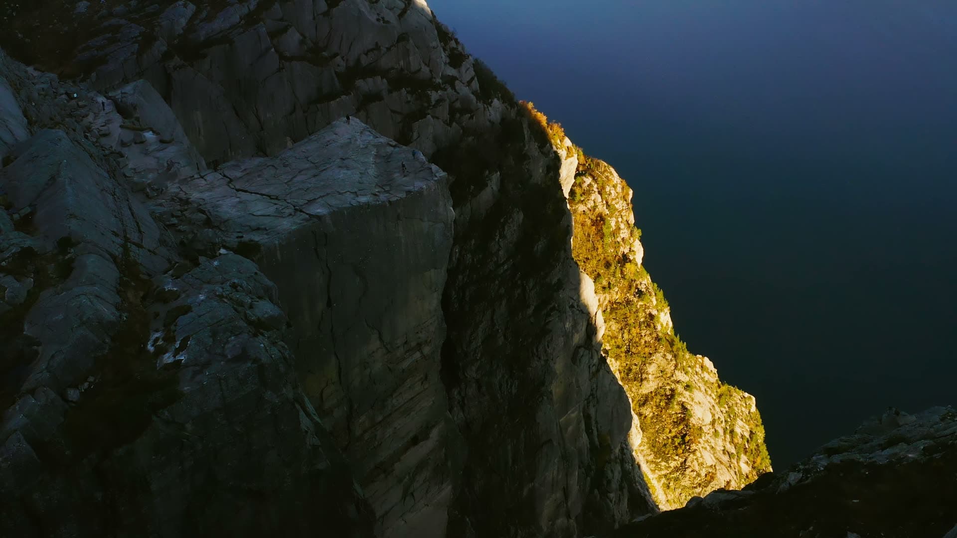 Sunlit moss-covered cliff face above dark water, Preikestolen, Norway